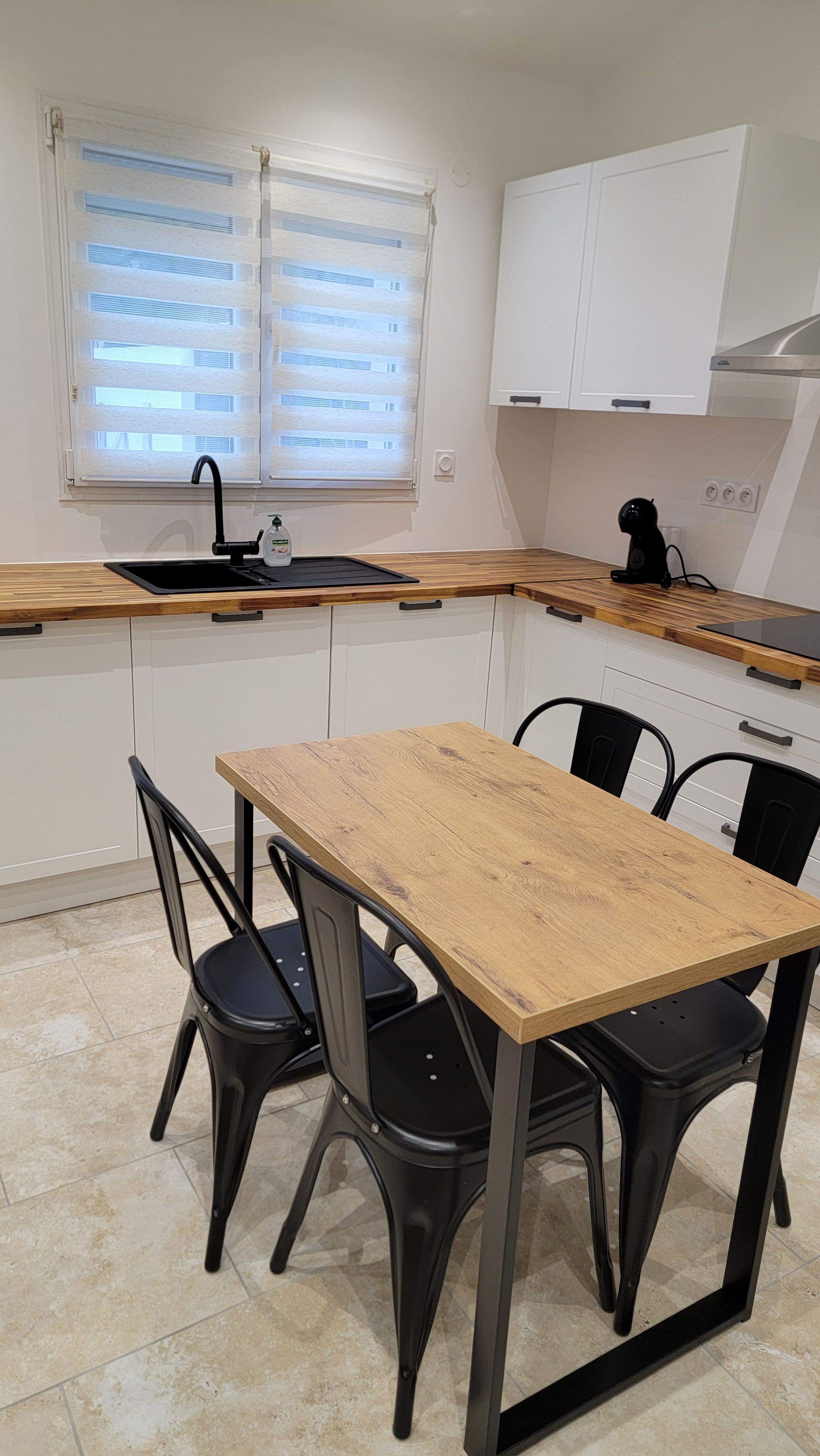 A compact kitchen with a wooden table and black chairs, a window with blinds, and white cabinets.