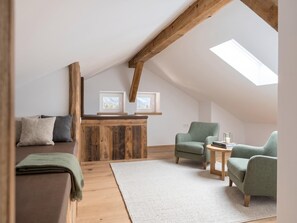 Quarto loft aconchegante com teto inclinado, vigas de madeira, claraboia, cama, duas poltronas, uma mesinha e um tapete.