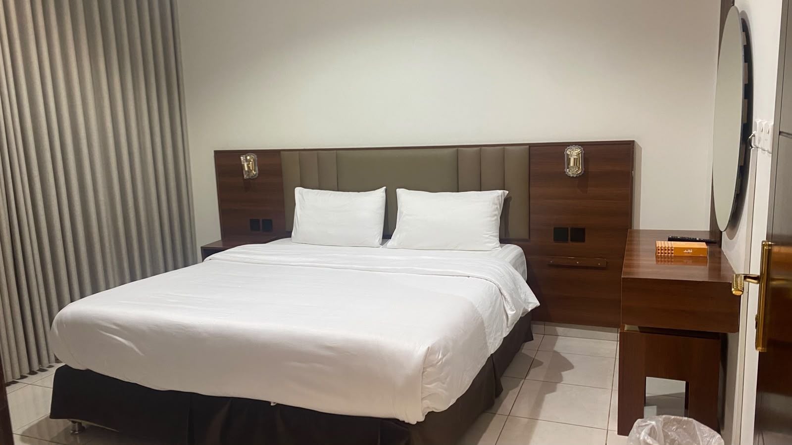 A hotel room with a bed, a wooden nightstand, a mirror, and a window with curtains.
