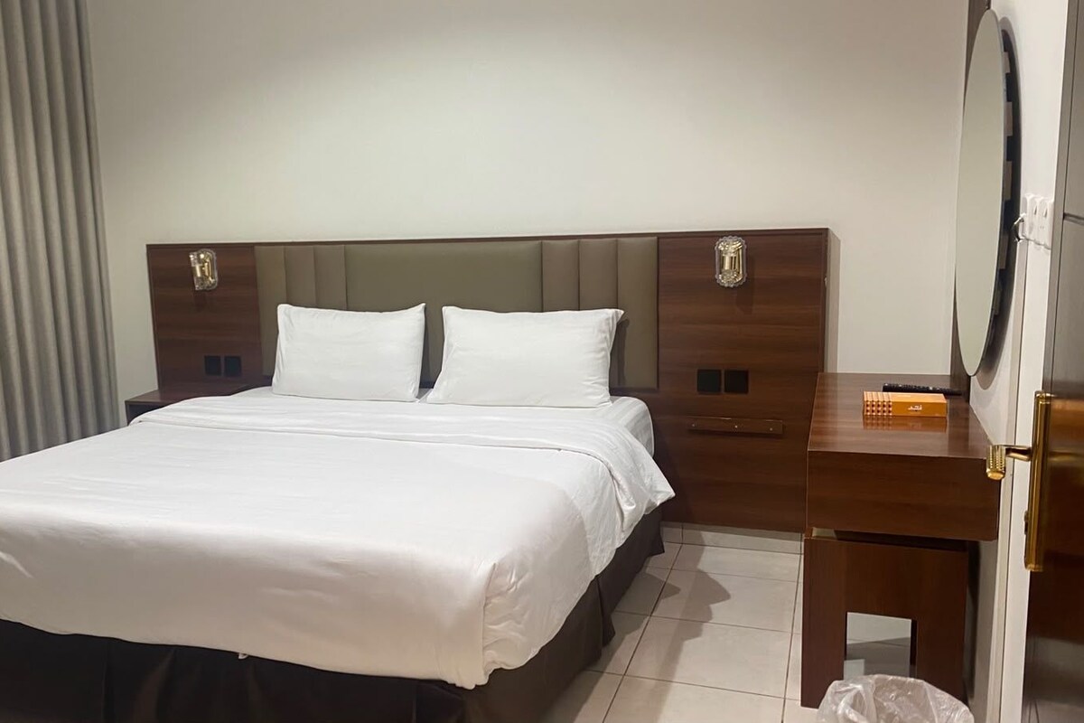 A hotel room with a bed, a wooden nightstand, a mirror, and a window with curtains.