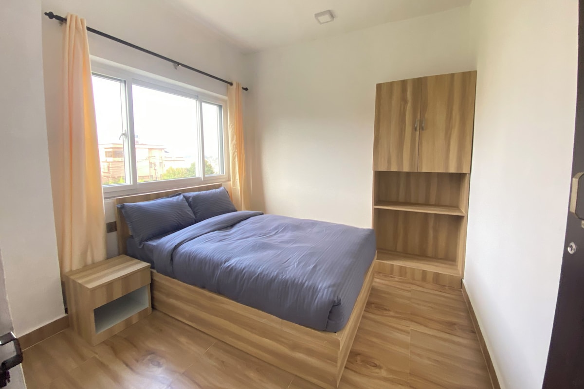 A bedroom with a bed, a window, a wooden wardrobe, and a small wooden table.