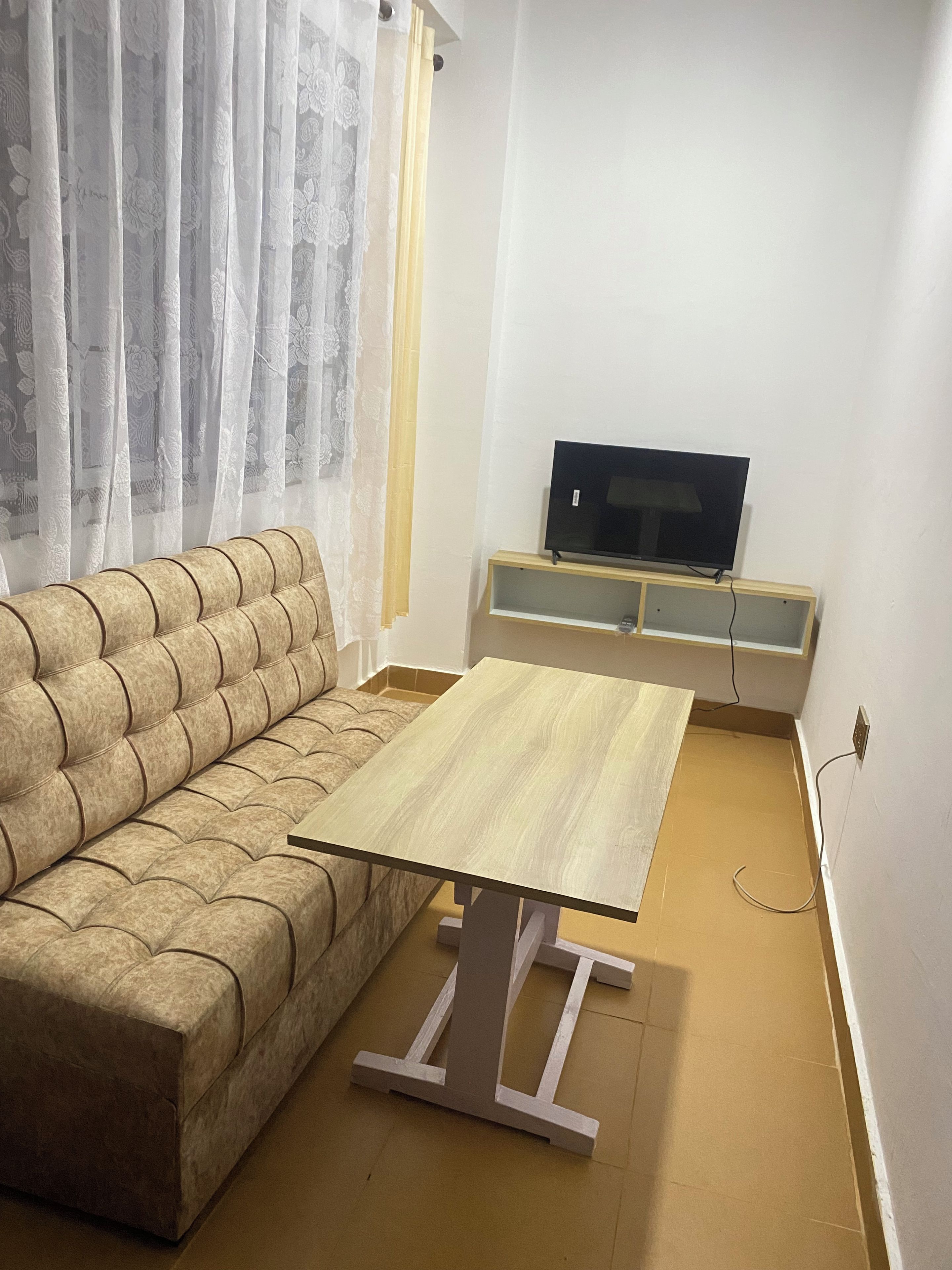 A living room with a sofa, a wooden coffee table, a TV on a stand, and a window with sheer curtains.