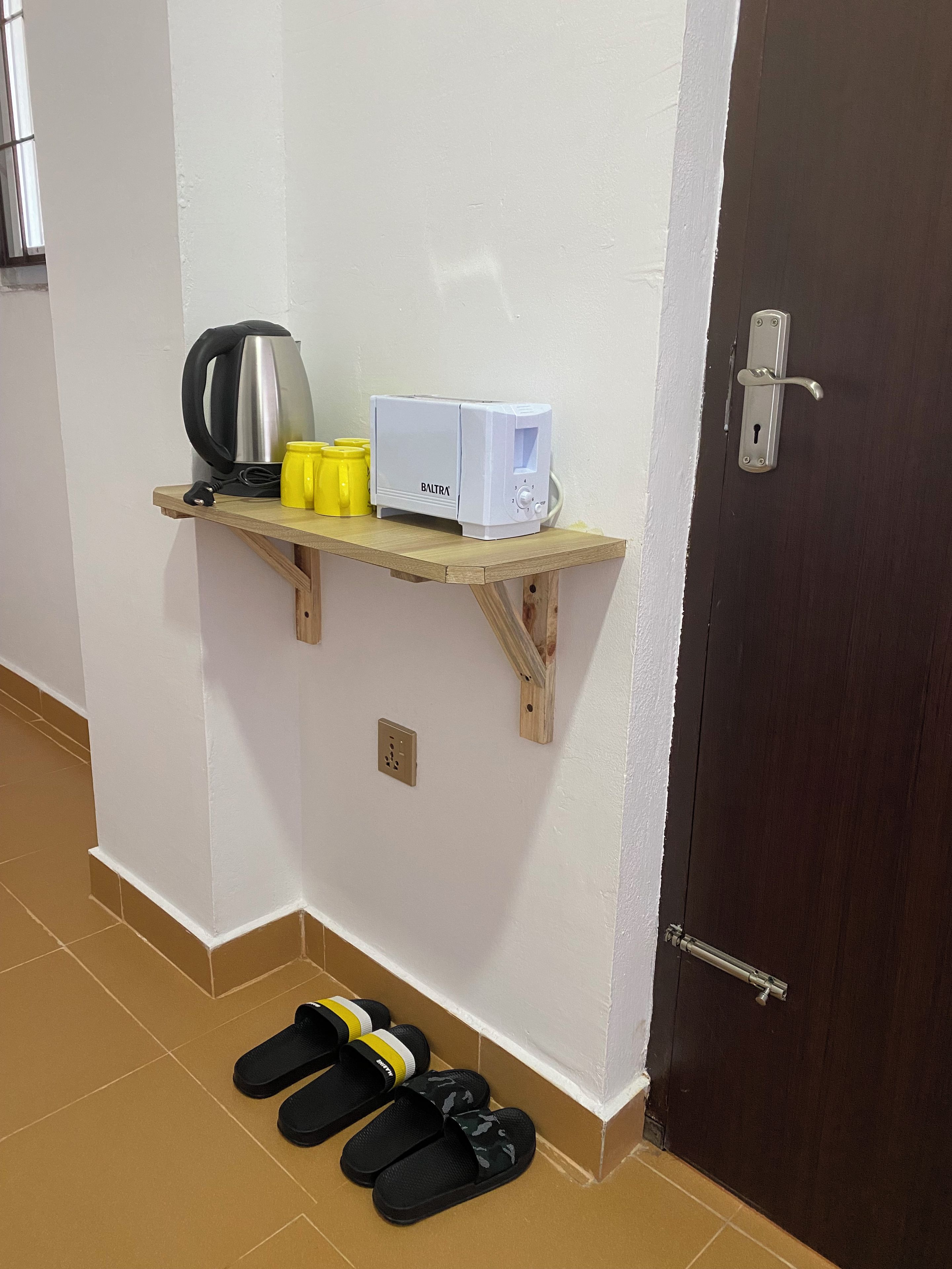 A small wooden shelf with a kettle, a toaster, and yellow cups against a white wall.