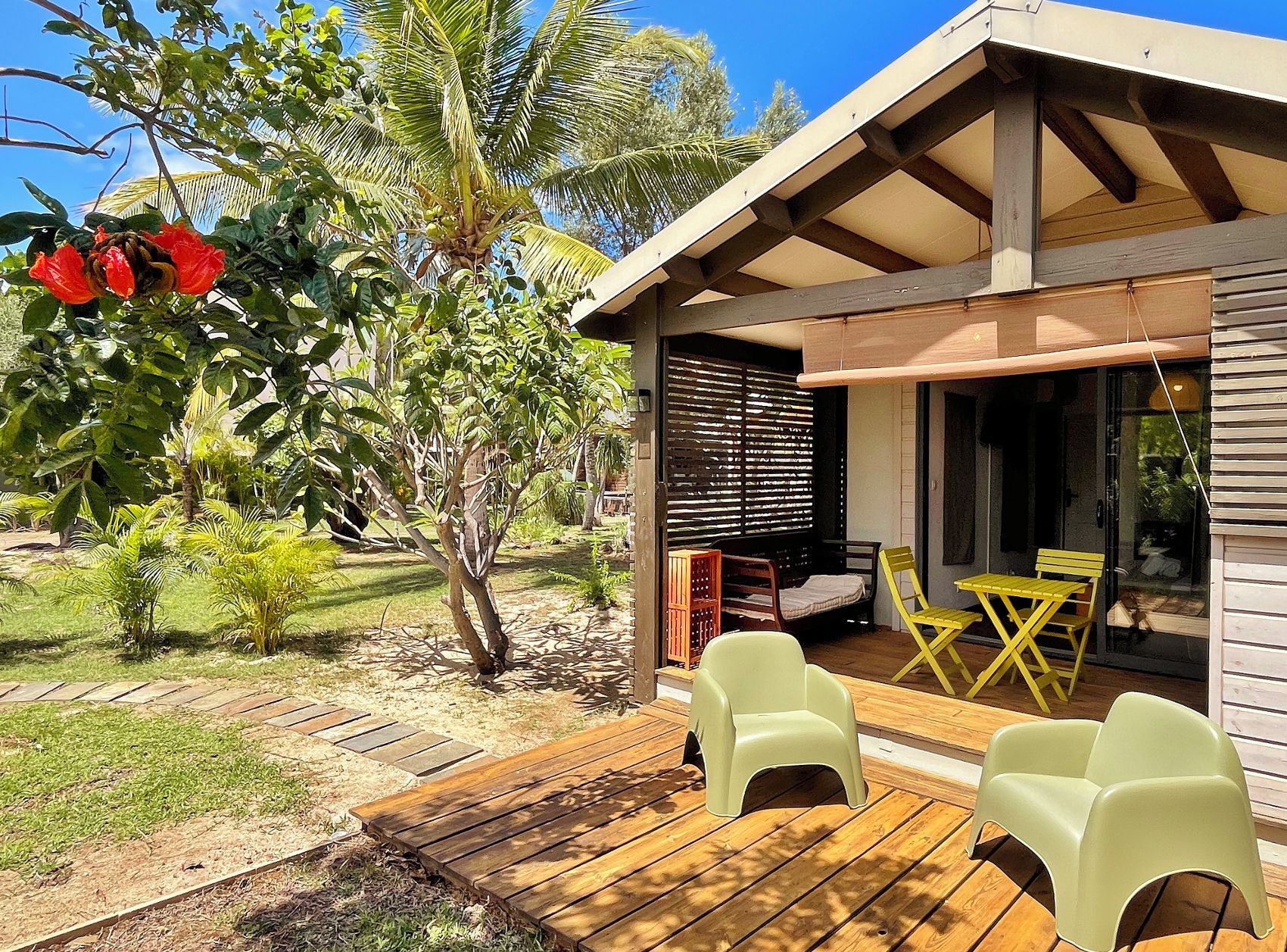 Una terraza de madera con mobiliario exterior, un espacio de estar techado y vistas a un jardín con palmeras y una flor roja.