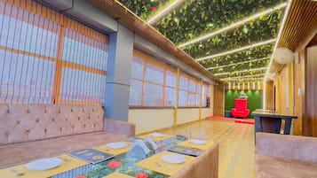 A restaurant with a long communal table, cushioned seating, and a green leafy ceiling.