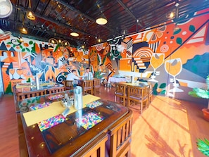 Un comedor interior con mesas y sillas de madera, un separador de vidrio con una etiqueta de nombre y un mural colorido en las paredes.