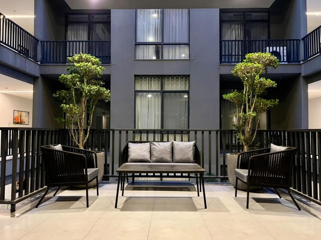 A modern interior with a sofa and chairs in a spacious area, a balcony with railings, and a building facade with windows and plants.