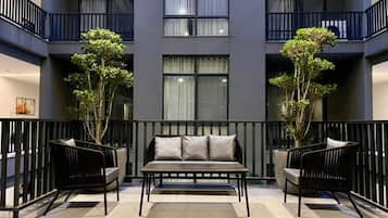 A modern interior with a sofa and chairs in a spacious area, a balcony with railings, and a building facade with windows and plants.