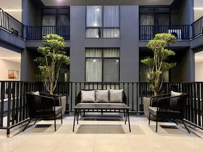Um interior moderno com sofá e poltronas em um espaço amplo, uma varanda com guarda-corpo e fachada de edifício com janelas e plantas.