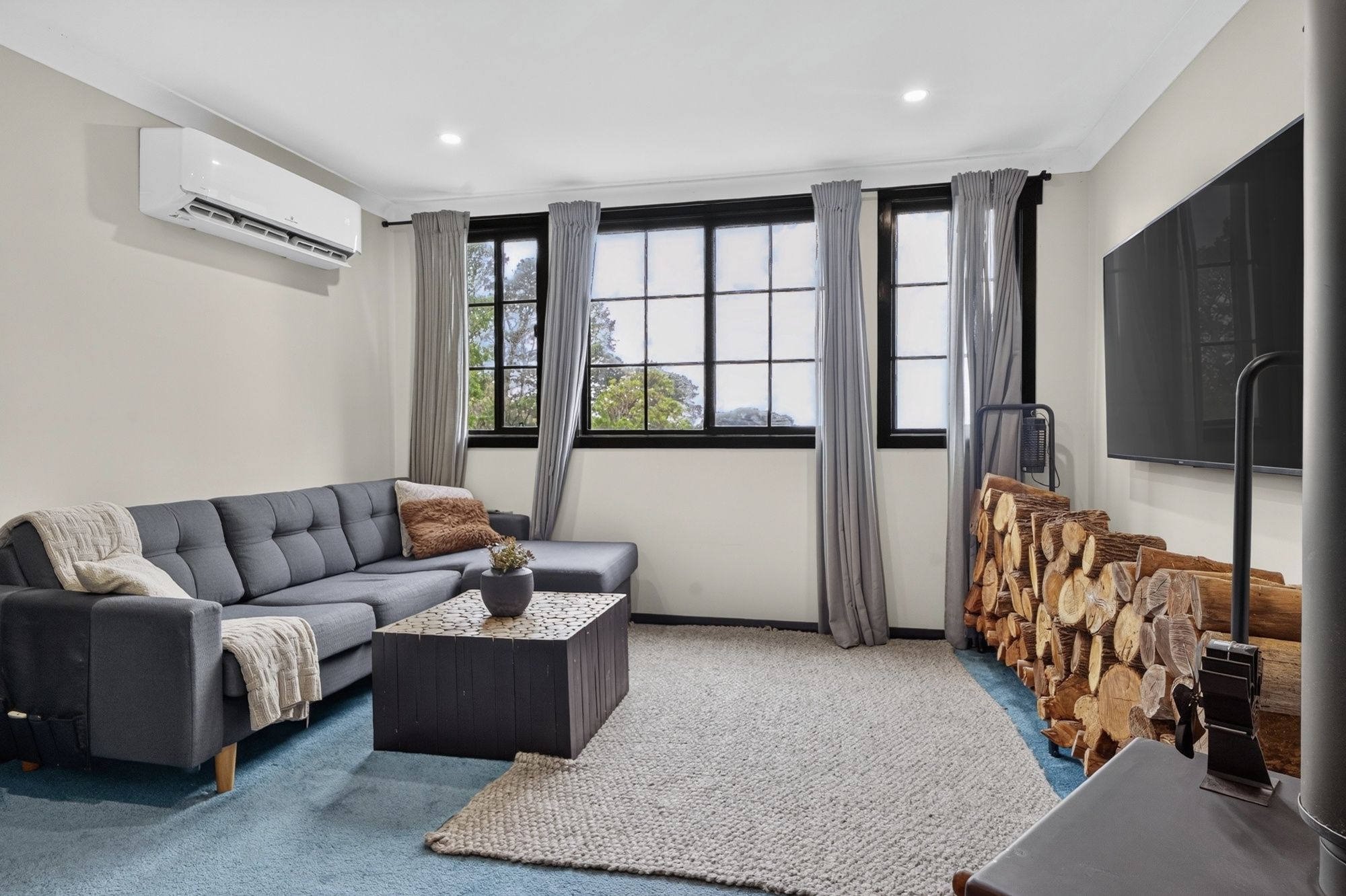 Una sala de estar moderna con un sofá seccional gris, una mesa de centro, un ventanal con cortinas, una chimenea con leña y un televisor montado en la pared.
