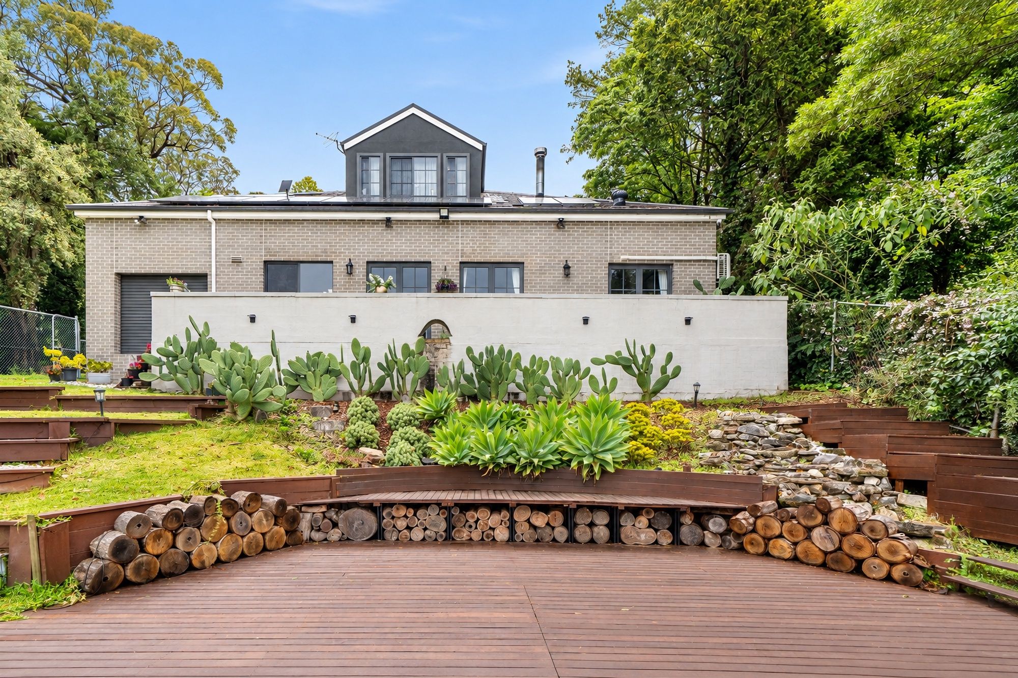 A modern two-story house with a flat roof, a chimney, and a garden with various plants and a wooden deck.