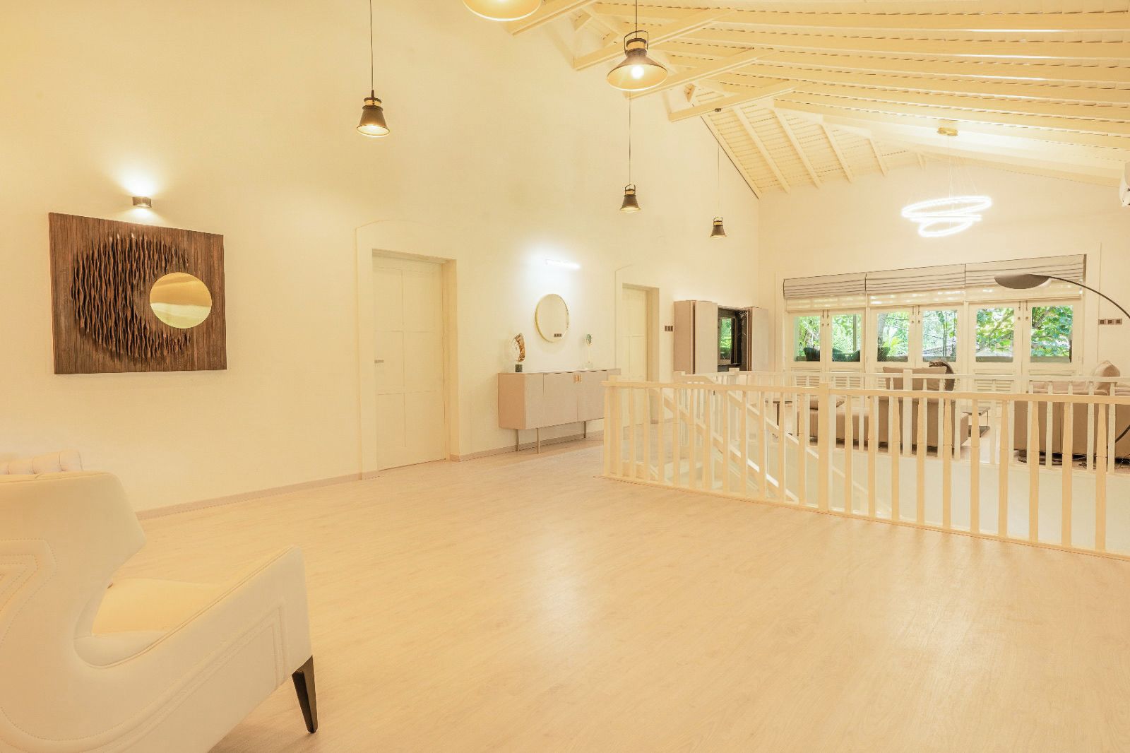 Quarto espaçoso com teto alto, sofá branco, parede de destaque em madeira e vista para o verde através de grandes janelas.
