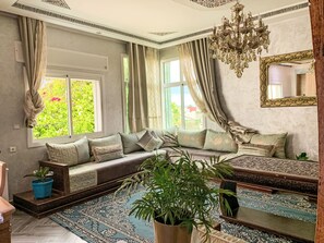 A room with a sofa set, a chandelier, a large mirror, and a view of greenery outside. - Jbila Maison by Interhome (Tanger)