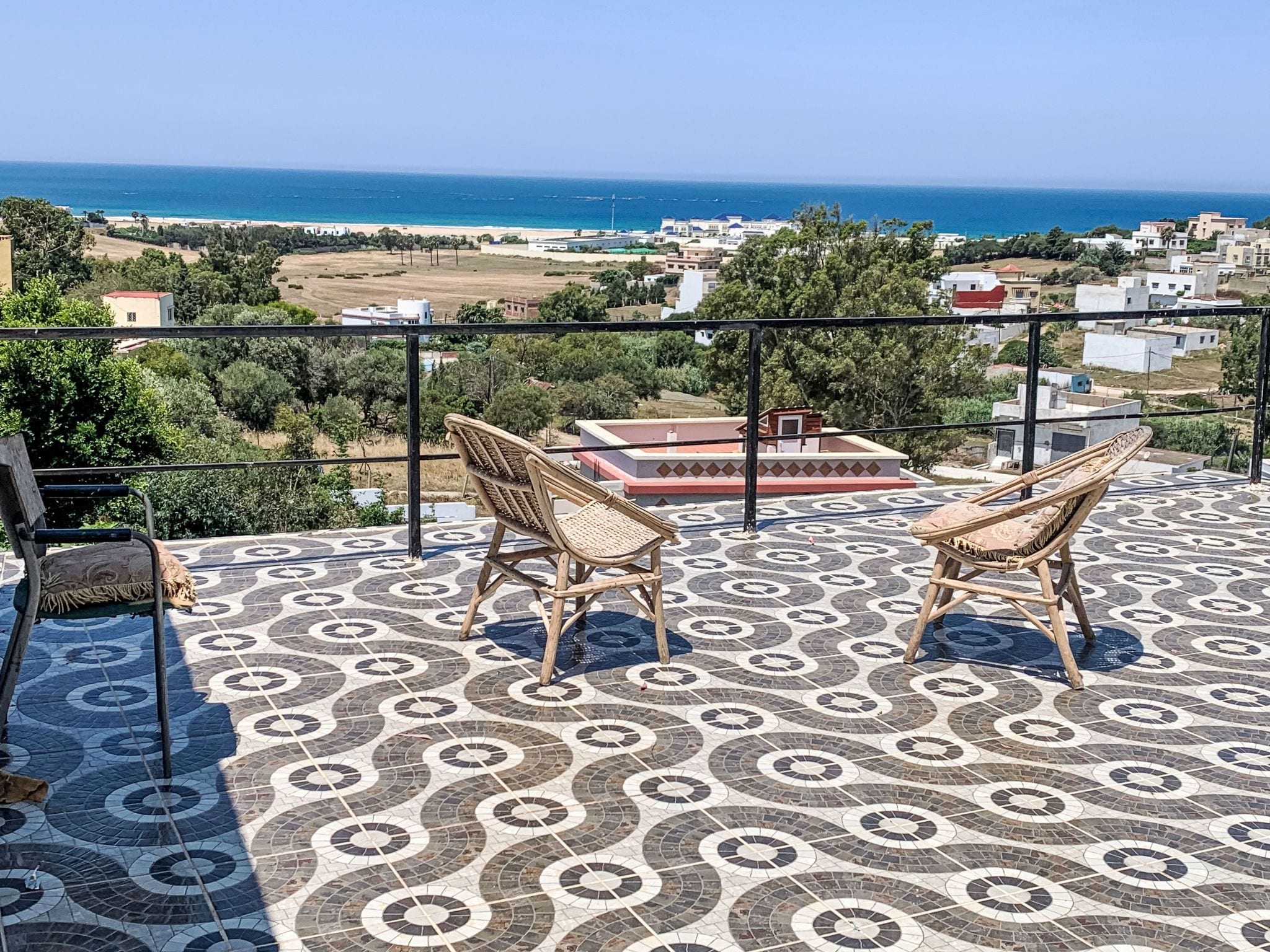 A terrace with two wicker chairs, a patterned floor, and a view of the sea and buildings in the distance.