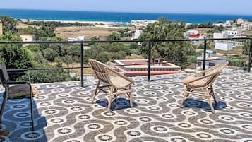 A terrace with two wicker chairs, a patterned floor, and a view of the sea and buildings in the distance.