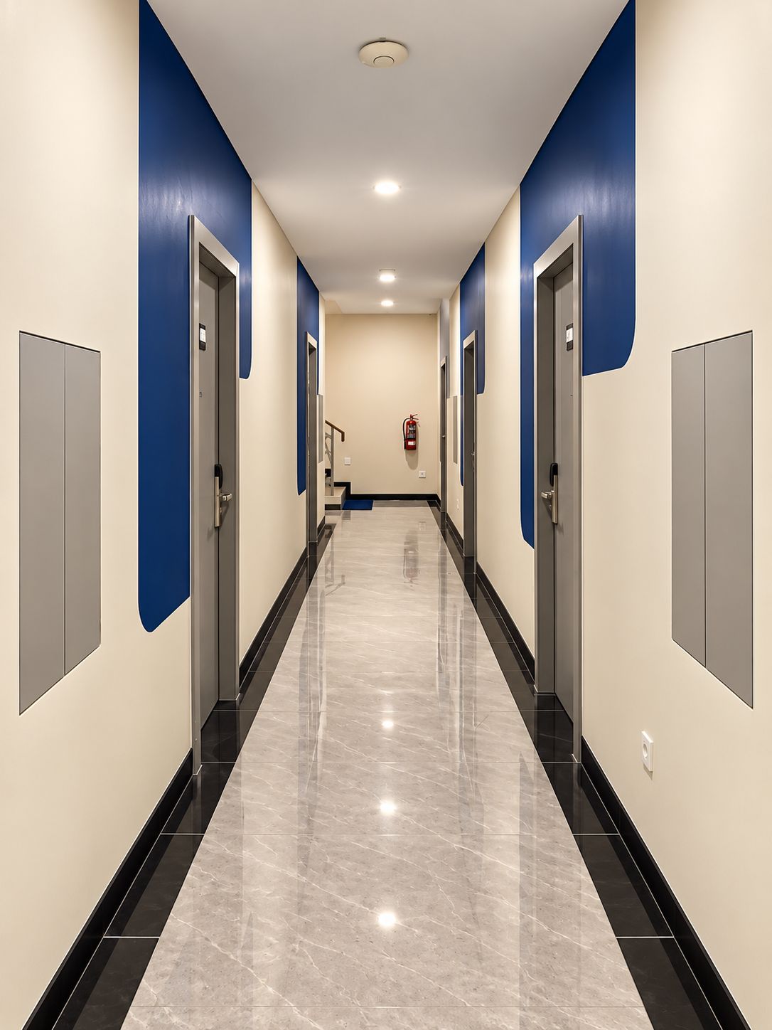 Un couloir avec des portes de part et d’autre, un extincteur fixé au mur et un luminaire au plafond.