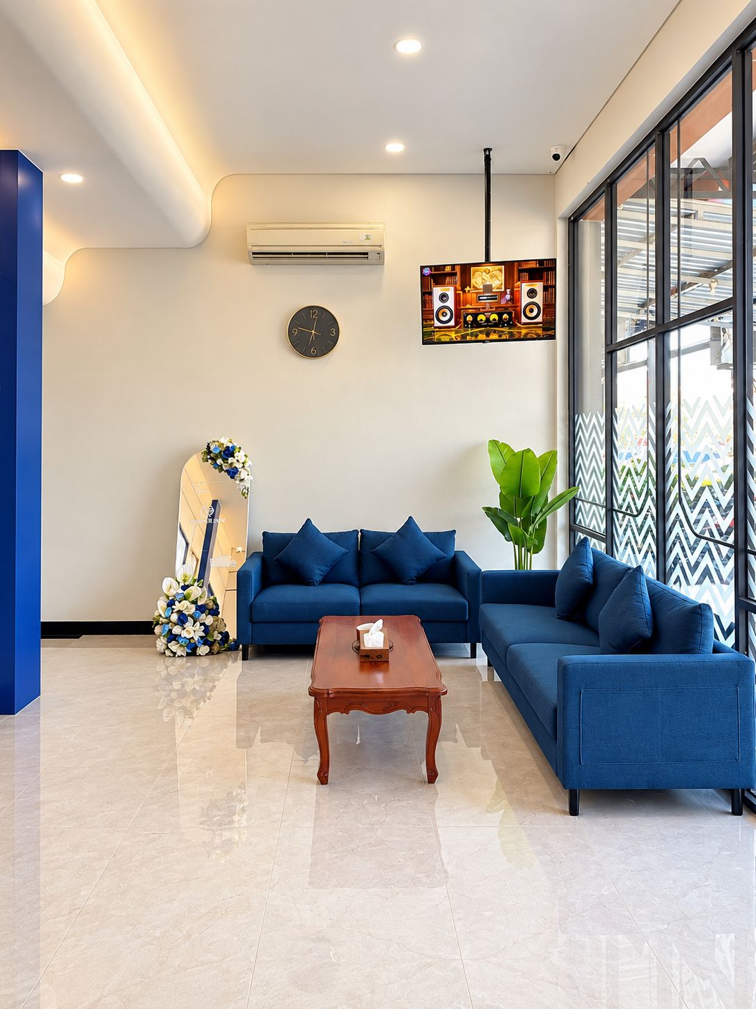 A modern living room with a blue sofa set, a wooden coffee table, a wall clock, and a large window with a view.