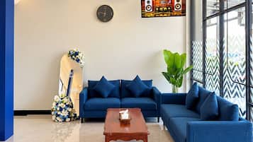 A modern living room with a blue sofa set, a wooden coffee table, a wall clock, and a large window with a view.