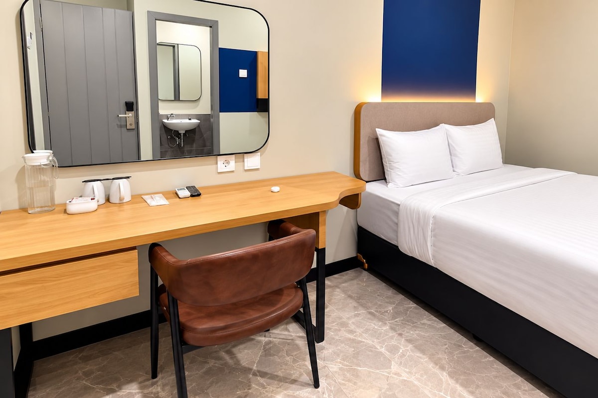 A hotel room with a bed, a desk, a chair, a mirror, and a nightstand.