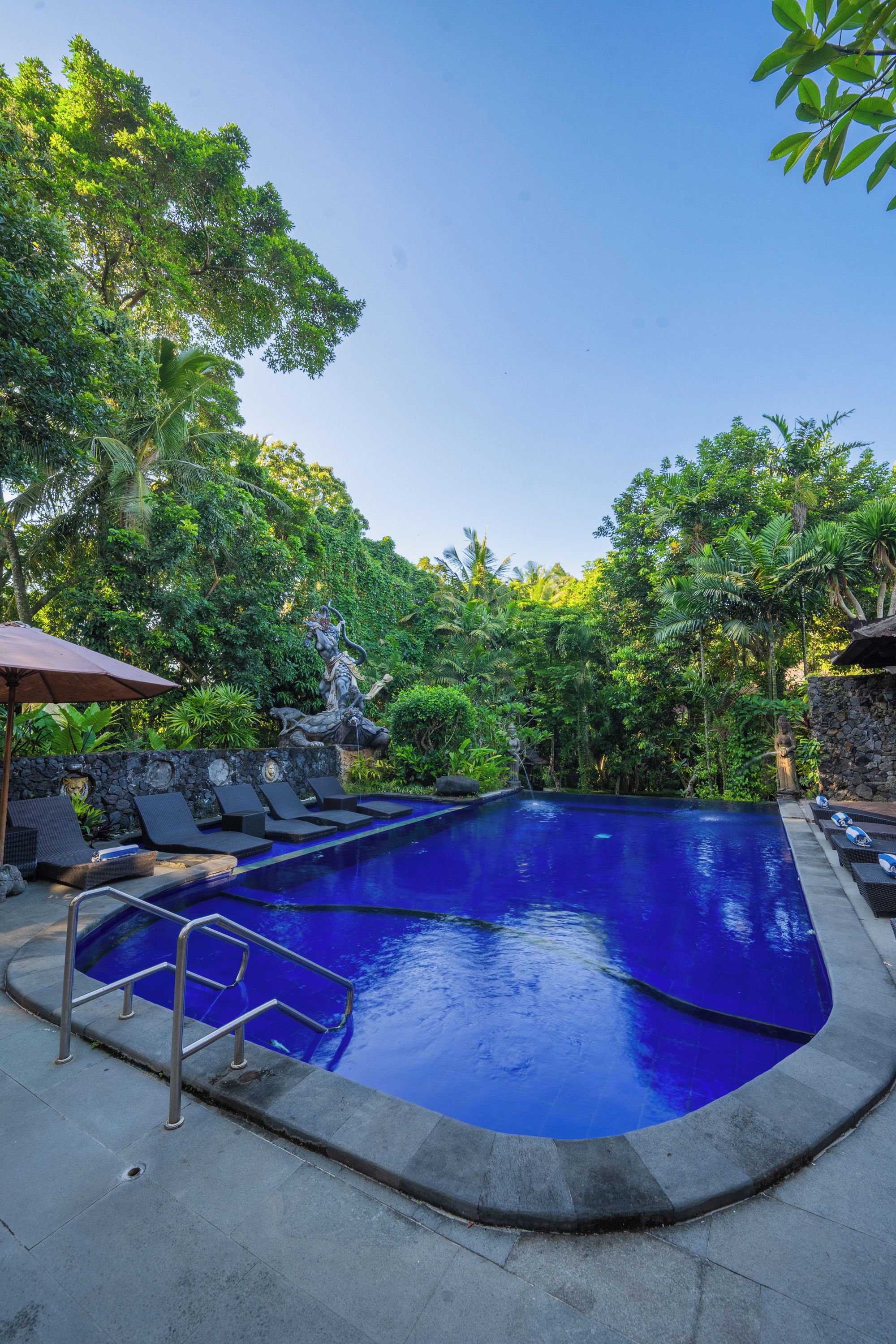 A swimming pool with a stone wall, surrounded by lush greenery and a statue.