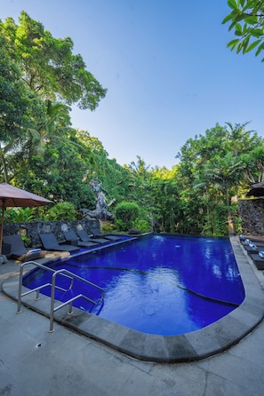Una piscina con una pared de piedra, rodeada de abundante vegetación y una estatua.