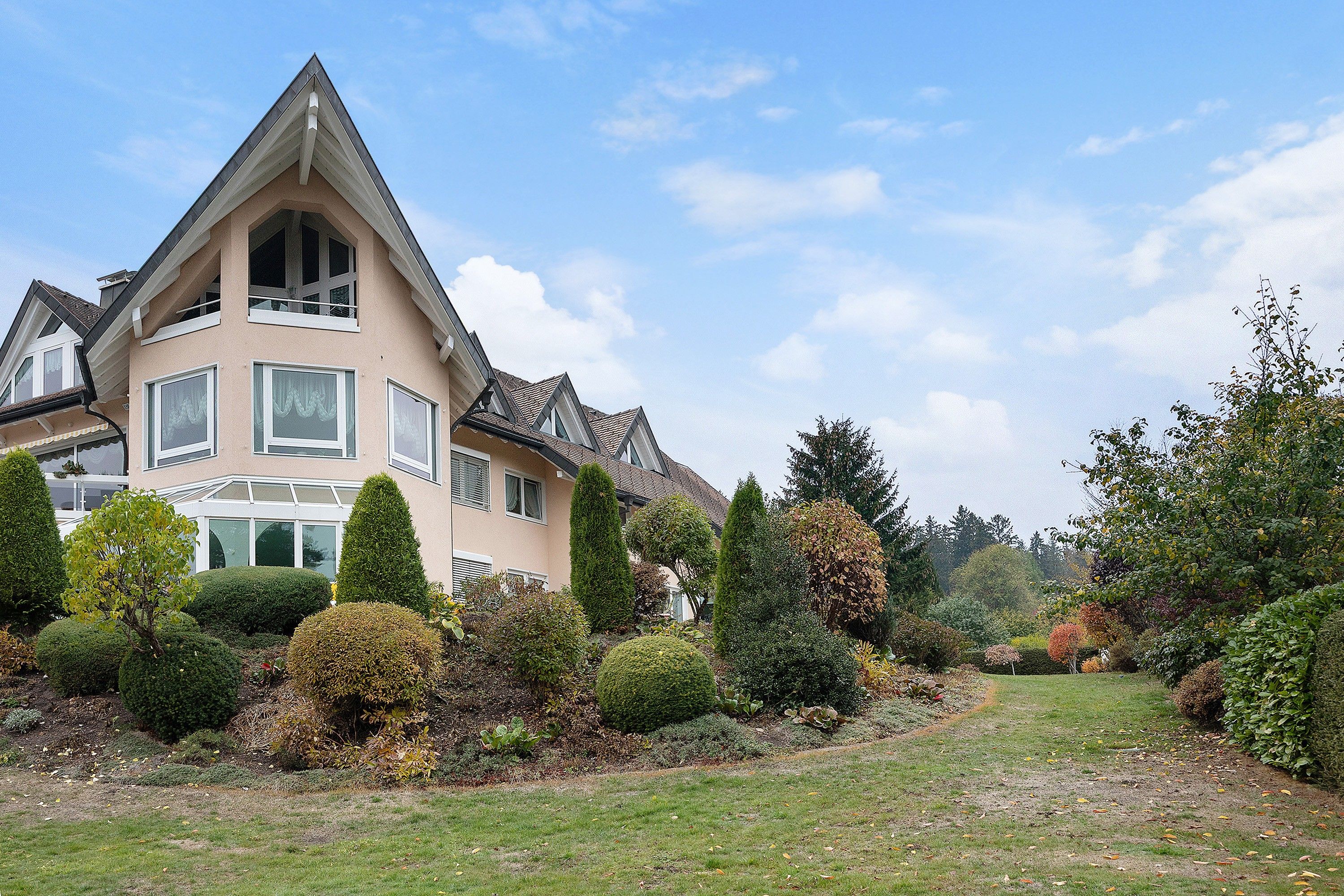 Ein großes, mehrgeschossiges Haus mit einem markanten Satteldach und einem gepflegten Garten mit verschiedenen Sträuchern und einer Rasenfläche.