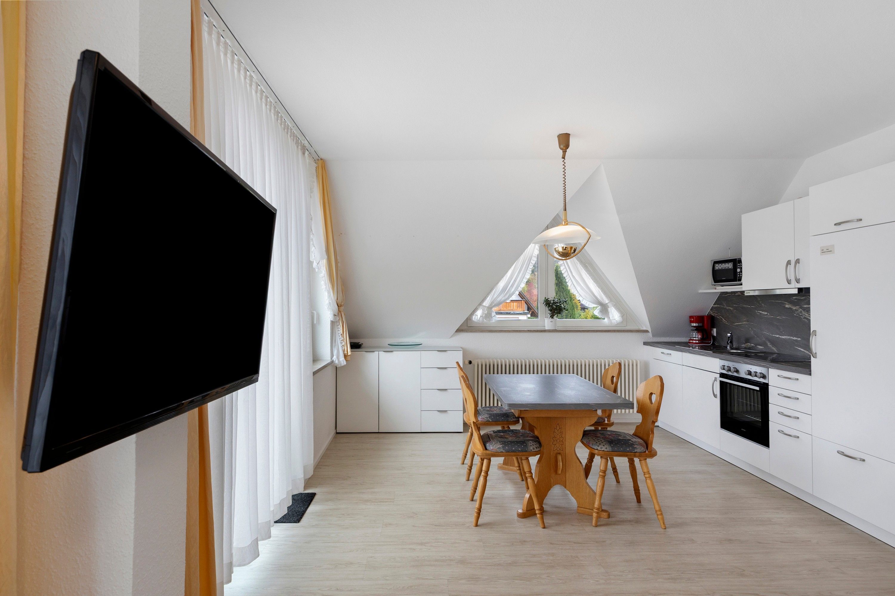 Eine moderne Küche mit weißen Schränken, einem Esstisch mit Stühlen, einem wandmontierten Flachbildfernseher und einem Fenster mit Vorhängen.