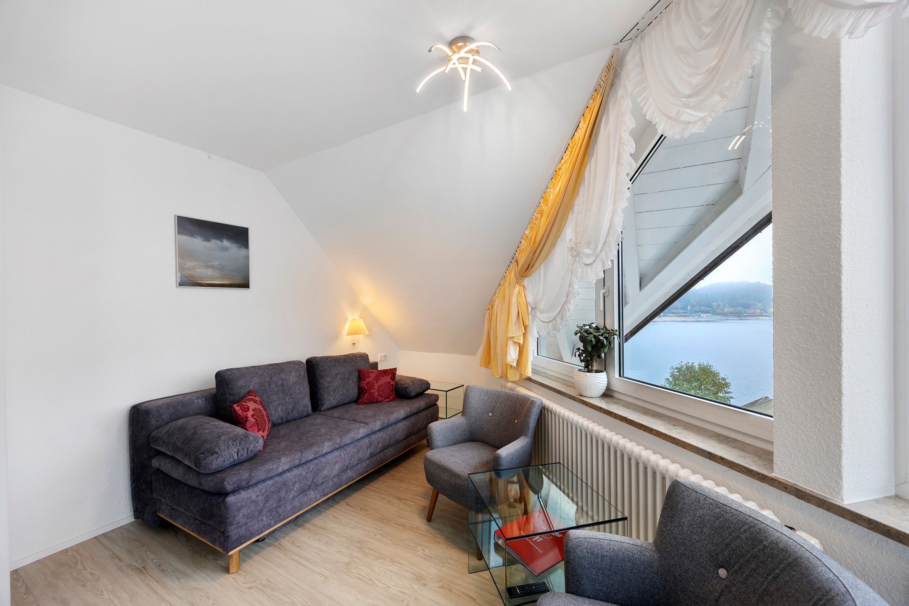 Ein modernes Wohnzimmer mit einem grauen Sofa, einem Glastisch und Blick auf ein Gewässer durch ein großes Fenster.