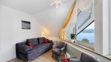 Ein modernes Wohnzimmer mit einem grauen Sofa, einem Glastisch und Blick auf ein Gewässer durch ein großes Fenster.