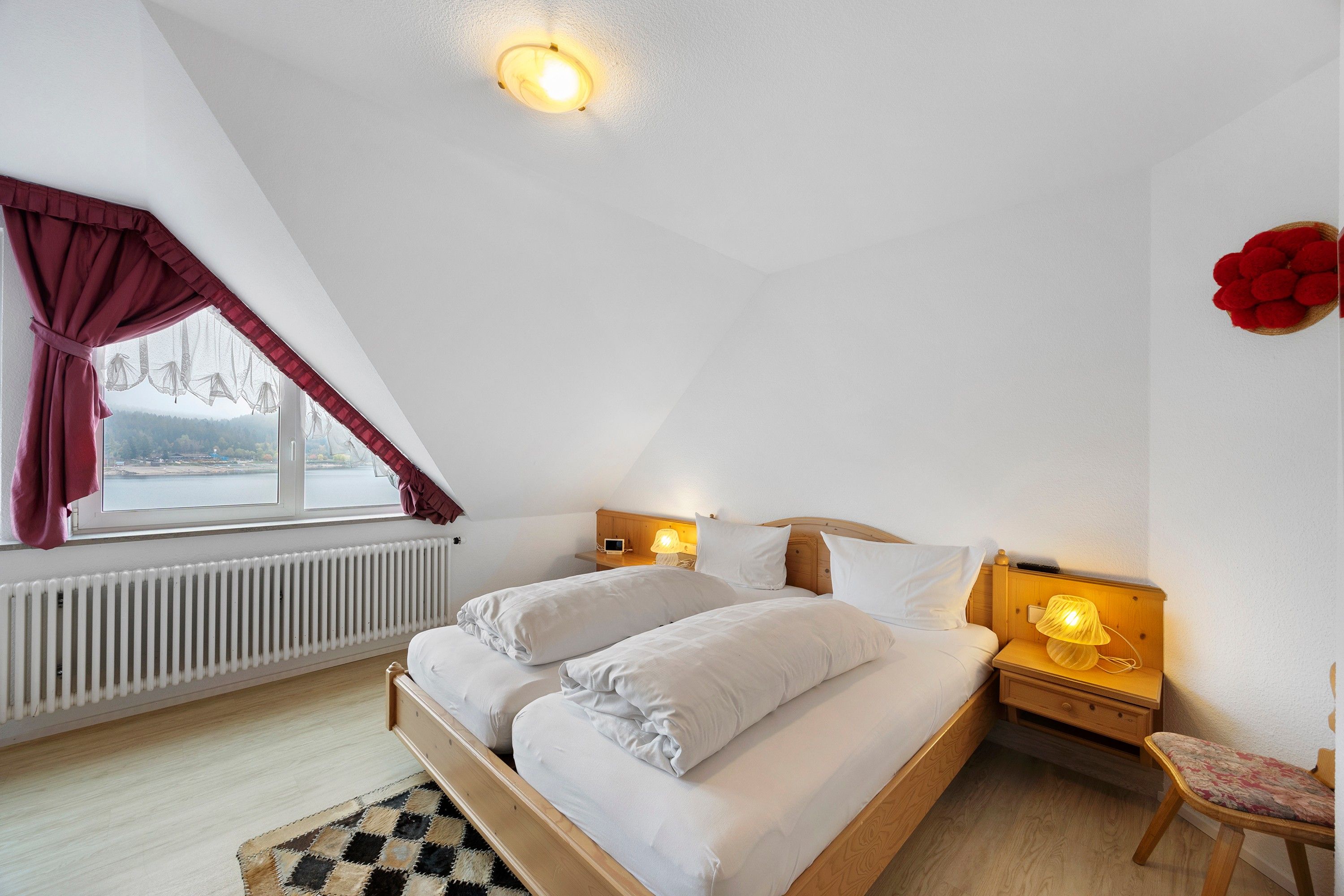 Ein Schlafzimmer mit Bett, Nachttisch, Sessel, einem Fenster mit Vorhängen und Blick ins Freie.