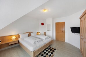 Une chambre avec un plafond en pente, un lit recouvert de draps blancs, une table de chevet avec une lampe, une armoire en bois et un téléviseur fixé au mur.