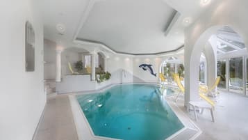 Ein Hallenschwimmbad mit klarem Wasser, umgeben von weißen Wänden und Säulen, sowie eine Sitzgelegenheit mit gelben Stühlen.