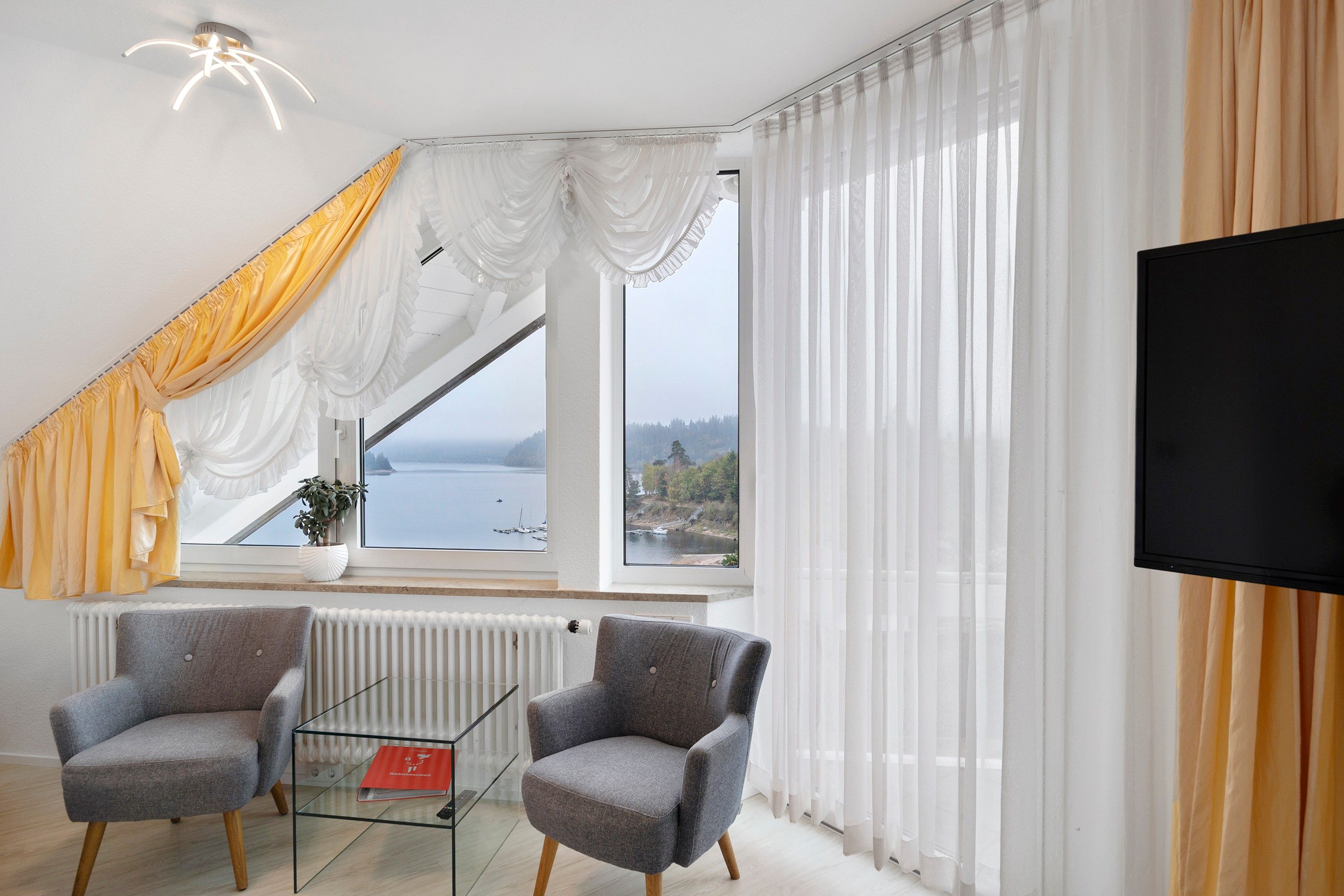 Ein modernes Wohnzimmer mit großem Fenster, zwei Sesseln, einem Glastisch und einem Flachbildfernseher.