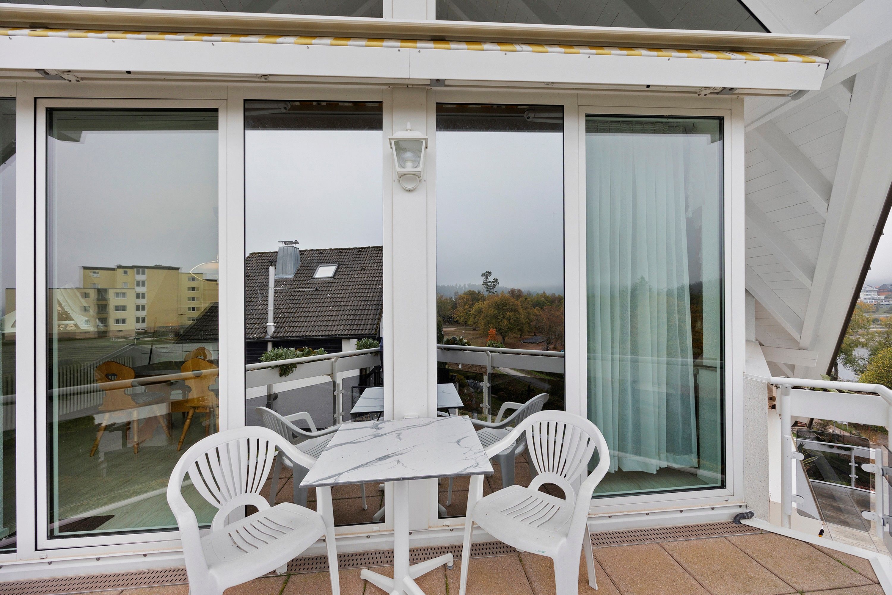 Ein Balkon mit Tisch und Stühlen, großen Fenstern und Blick auf Gebäude und Bäume.