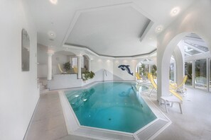 Una piscina interior con agua cristalina, rodeada de paredes y columnas blancas, y una zona de estar con sillas amarillas.
