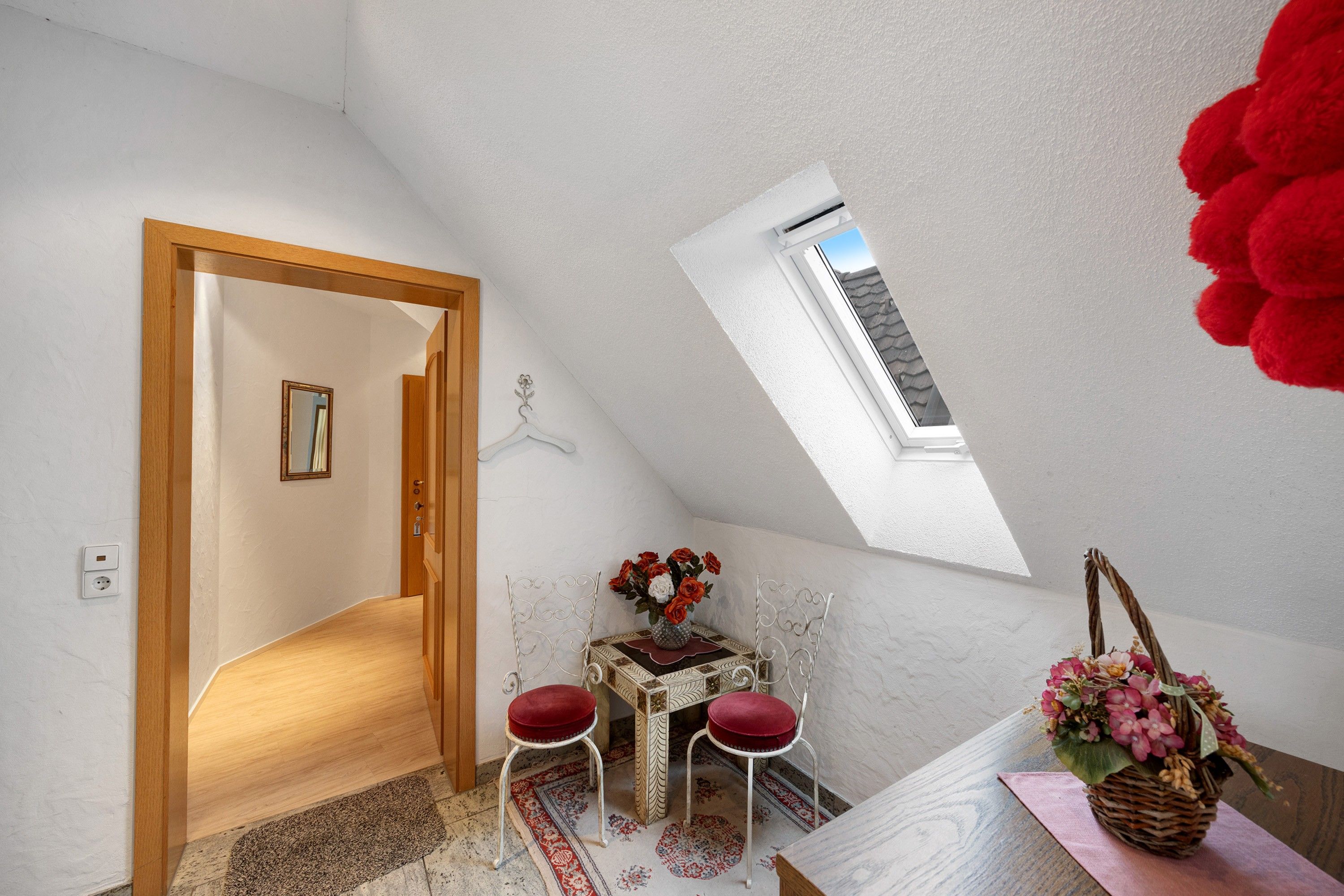 Een kamer met een schuine plafond, een klein tafeltje met stoelen, een spiegel en een raam.