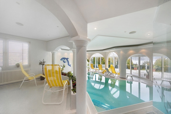 Une piscine intérieure à l’eau claire, entourée de chaises longues jaunes et de colonnes blanches.