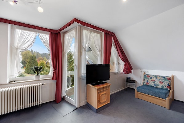 Una habitación con televisión, un pequeño armario de madera, un banco con cojín floral y una ventana con cortinas.