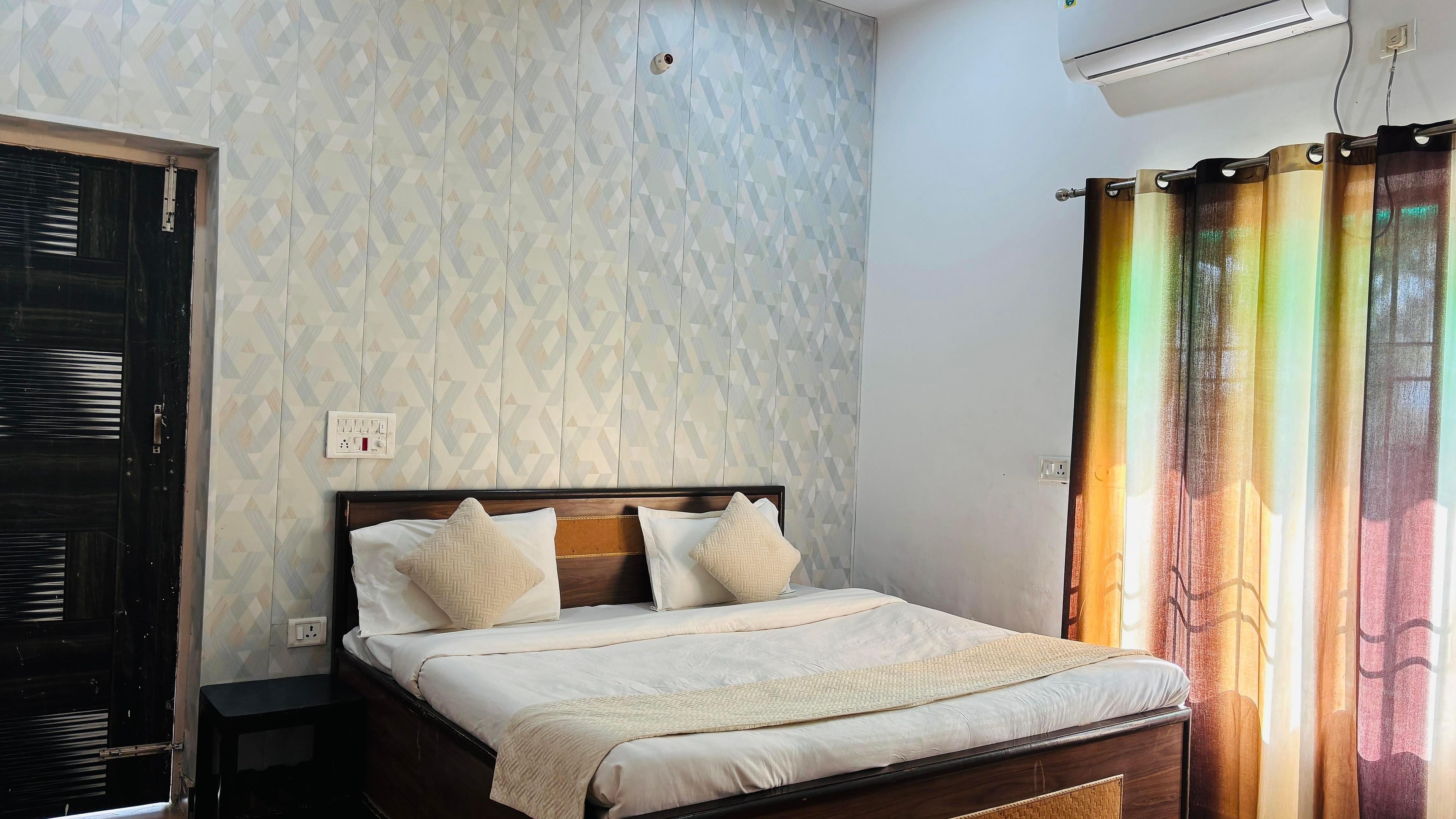 A bedroom with a bed, a nightstand, a wall socket, and a window with curtains.