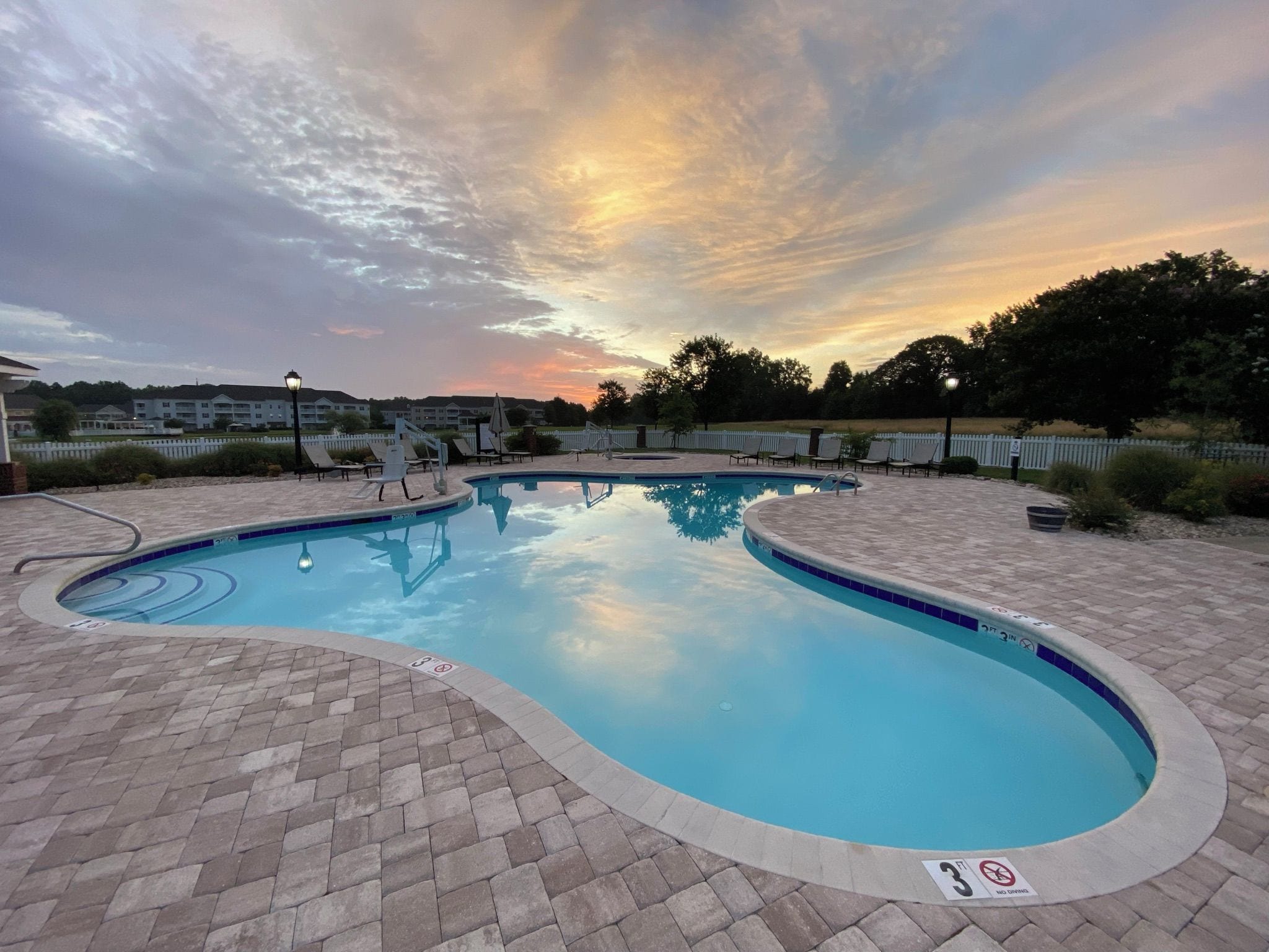A swimming pool with a brick deck, surrounded by lounge chairs and a white fence.