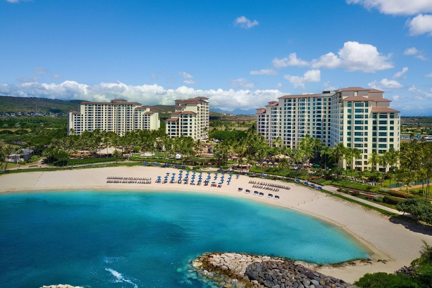 Un hotel frente a la playa con varios edificios altos, una playa arenosa con sombrillas y un océano azul y cristalino.