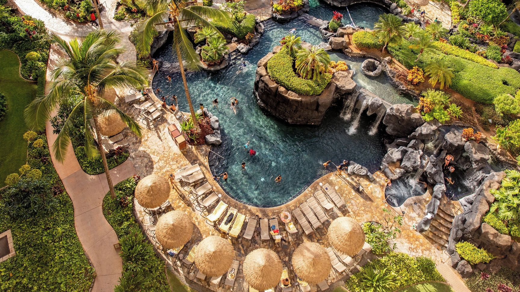 Una piscina de resort con múltiples elementos acuáticos, incluyendo cascadas y una zona de piscina central rodeada de sillones y sombrillas.