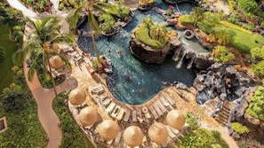 Una piscina de resort con múltiples elementos acuáticos, incluyendo cascadas y una zona de piscina central rodeada de sillones y sombrillas.