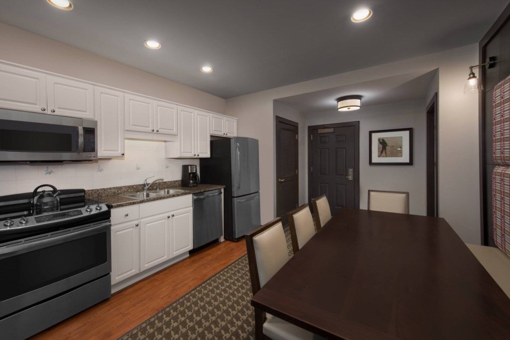 A modern kitchen with white cabinets, a stainless steel microwave, a built-in oven, and a dark refrigerator. There is a dining table with chairs and a patterned rug.