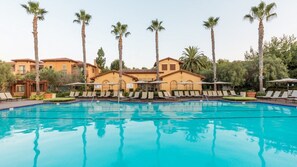 Ein Swimmingpool mit Liegestühlen und Palmen, umgeben von Gebäuden im mediterranen Stil.