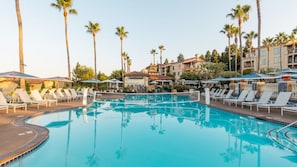 En swimmingpool med solstolar och parasoller, omgiven av palmer och hotellbyggnader.