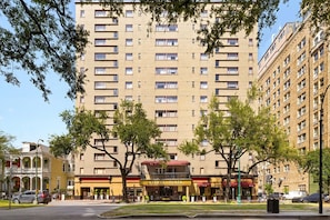 Um edifício de vários andares com entrada central, rodeado por árvores e uma rua.