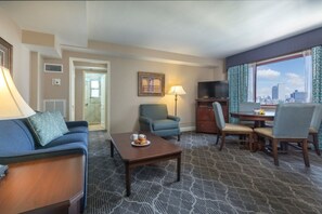 Quarto de hotel com sofá, poltrona, mesa de jantar, televisão e vista para a cidade.