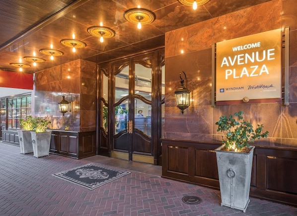 En hotellentrén med en skylt som lyder ”VÄLKOMMEN TILL AVENUE PLAZA”, en entré med glasdörr och krukväxter.