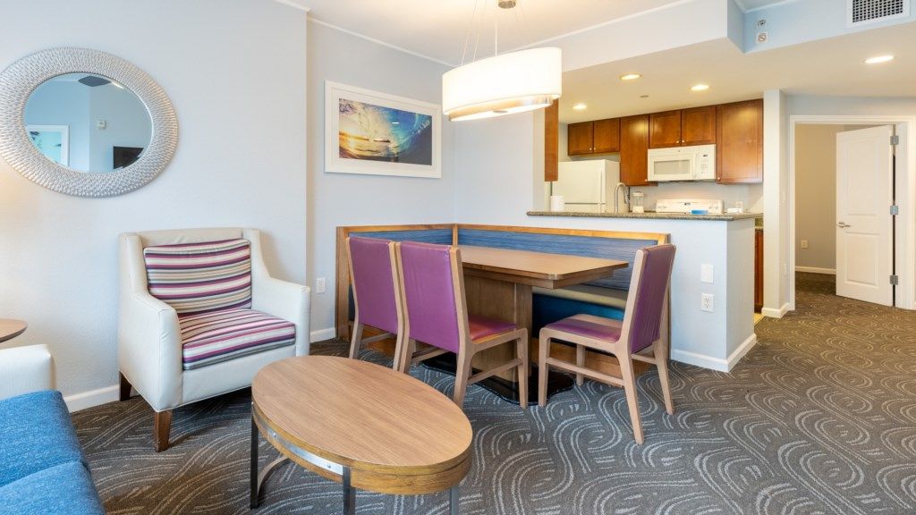 Une chambre d’hôtel compacte comprenant un coin cuisine, un espace repas avec une table et des chaises, un fauteuil à rayures et un miroir rond.
